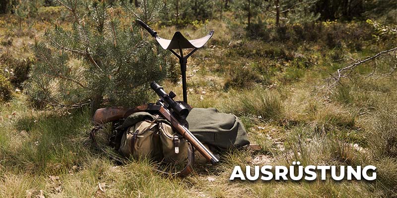 Jägerausrüstung liegt im grasigen Gelände: ein Rucksack, eine Decke, ein Gewehr mit Zielfernrohr und ein Klapphocker stehen neben einem kleinen Baum; unten rechts steht der Schriftzug ‚Ausrüstung‘.